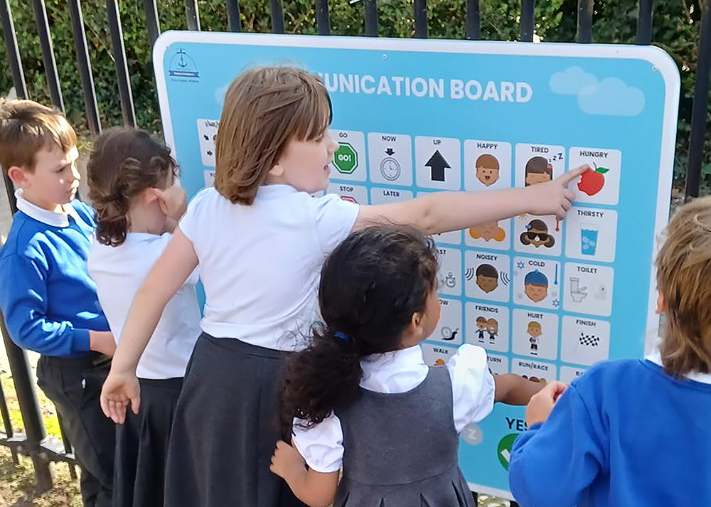 A Voice 4 All Primary School Communication Board mounted externally on a black rail fence with five primary school children,  communicating using the board with one girl pointing at an 'hungry' icon