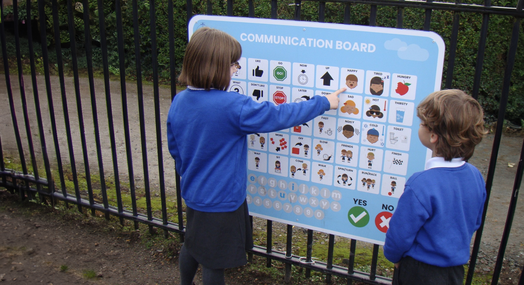 A Voice 4 All Primary School Communication Board mounted externally on a black rail fence with two primary school children, one male, one female communicating using the board with the girl point at an 'happy' icon